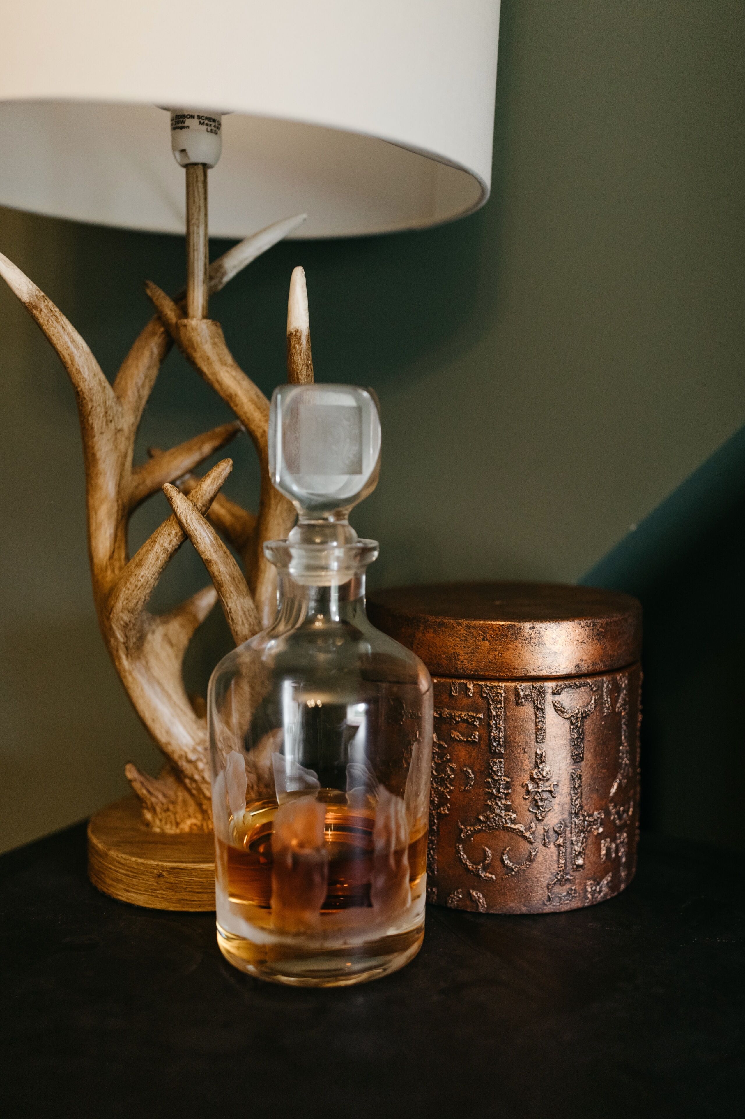 Una botella de vidrio transparente con un tapón, un recipiente de cobre con diseños en relieve, y una decoración de asta de ciervo sobre una superficie oscura.