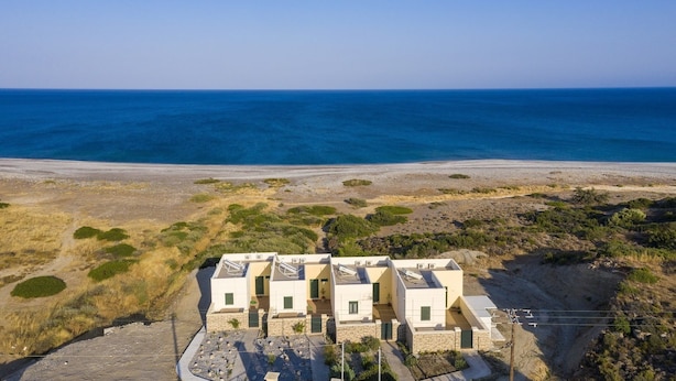 A row of modern, white, two-story buildings with flat roofs, situated on a rocky terrain with sparse vegetation, overlooking a wide, calm sea. - Holiday House Myrtos (Rhodes)