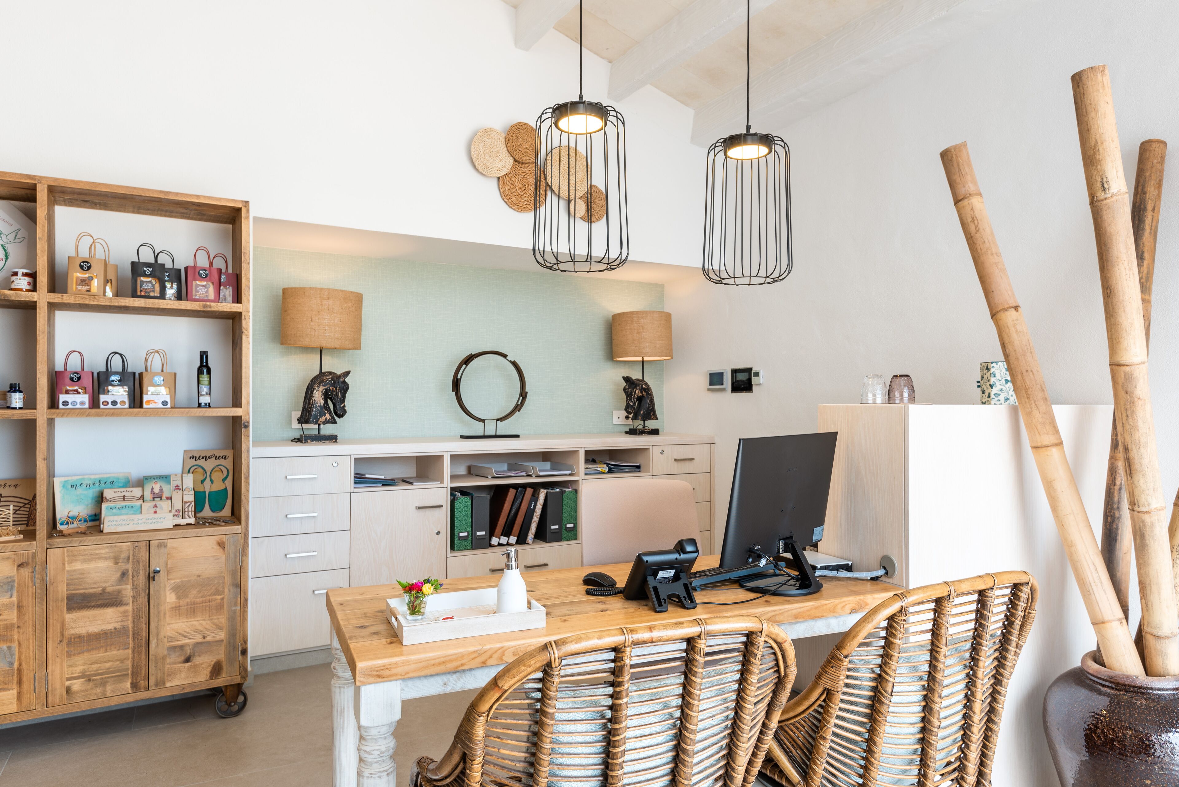 Een werkplek aan huis met een houten bureau, rieten stoelen, een computer, planken met tassen en een lamp.
