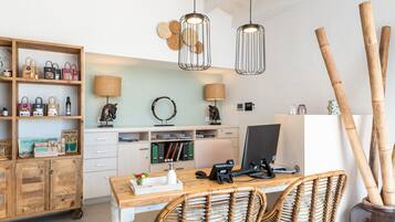 Een werkplek aan huis met een houten bureau, rieten stoelen, een computer, planken met tassen en een lamp.