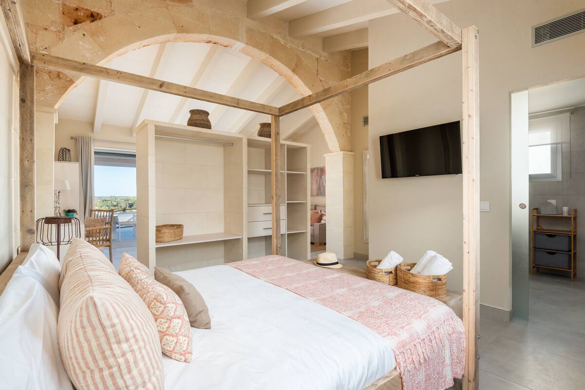 Ein Schlafzimmer mit einem großen Bett, Holzbalken, einem Fernseher und Blick ins Freie.
