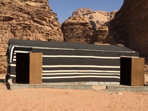 Uma pequena construção de um único cômodo, com fachada listrada, localizada em um ambiente desértico com penhascos rochosos ao fundo.