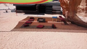 A desert setting with a seating area featuring red cushioned benches, a small table, and a fire pit. - Al sultan bedouin camp (Wadi Rum)