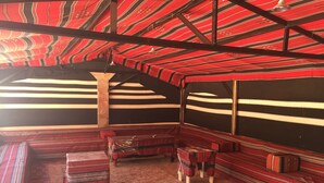 A room with a high ceiling, striped red and black walls, and a carpeted floor. There are benches and a small table with a chair. - Al sultan bedouin camp (Wadi Rum)