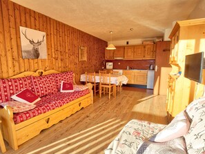 A cozy cabin interior with a wooden wall, a sofa with patterned cushions, a dining table with chairs, and a kitchen area with wooden cabinets and a television.