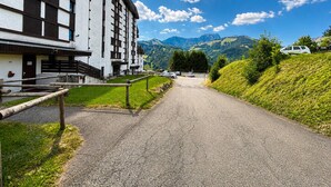 Ein Gebäude mit heller Fassade und dunklen Holzakzenten, ein gepflasterter Weg dorthin und eine gebirgige Landschaft im Hintergrund.