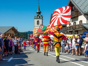 Un défilé avec des participants vêtus de costumes colorés et brandissant des drapeaux, une foule de spectateurs et, en arrière-plan, le clocher d’une église.