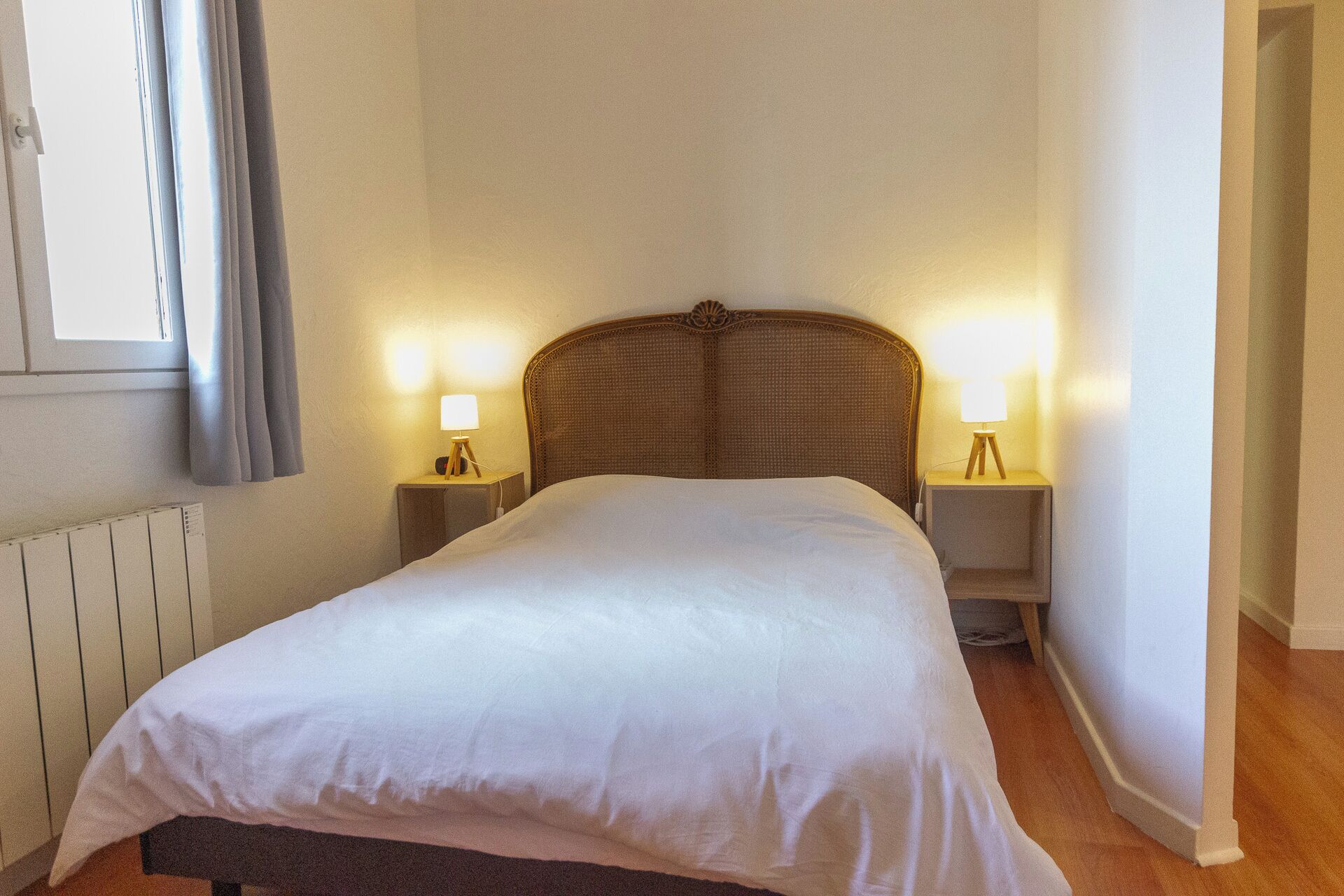 Ein ordentlich bezogenes Bett in einem Hotelzimmer, mit Kopfteil, zwei Nachttischlampen und einem mit Vorhängen versehenen Fenster.