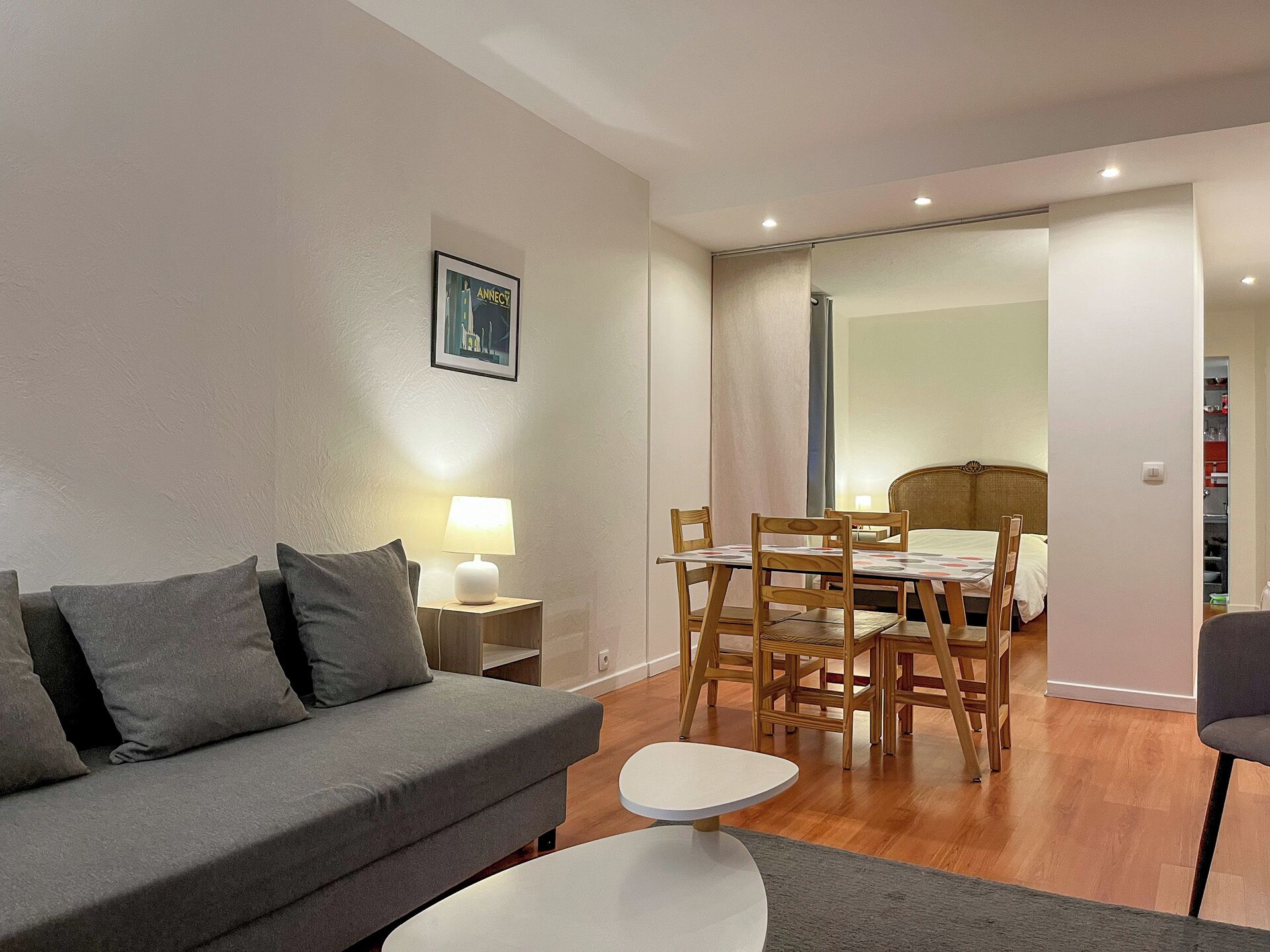 A modern living room with a grey sofa, a small round table, and a dining area with wooden chairs.