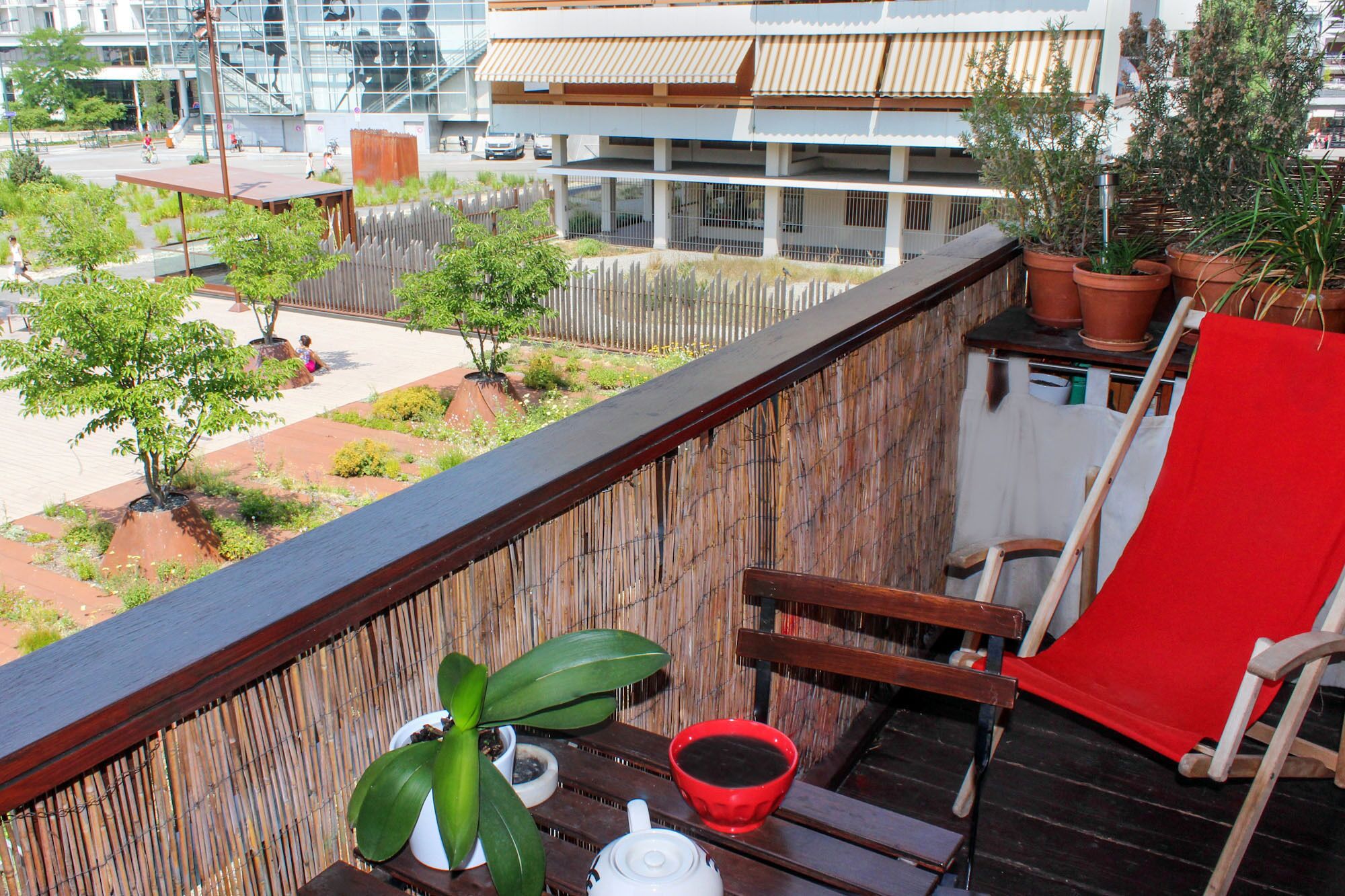 A balcony with a view of a garden, a red chair, a white teapot, and a red cup.