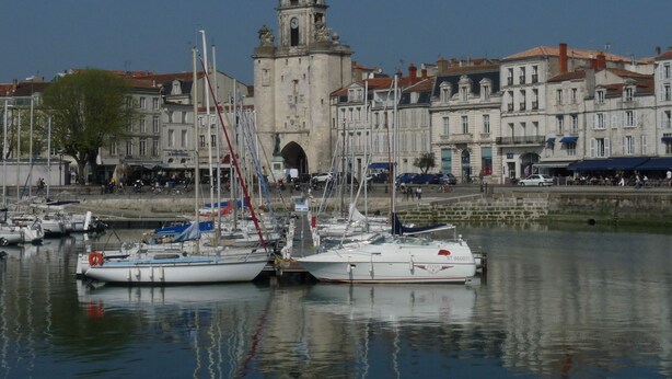 A marina with several sailboats docked, a historic building with a clock tower in the background, and a waterfront promenade with buildings and shops.