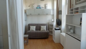 A small, well-lit room with a grey sofa, a small table, and a television. There are white cabinets above the sofa and a radiator to the right.