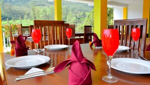 A dining table set with red napkins, white plates, and red wine glasses, overlooking a lush green landscape. - Grand Adams Peak Hotel (Nallathanniya)
