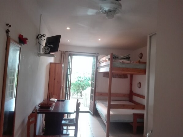 A compact room with a bunk bed, a small table, and a chair. There is a ceiling fan, a window with curtains, and a door leading outside. - Beach<50m.Equipped terrace. Parking. Car and bicycle options. Wifi. No. of activities (Marine de Bravone)