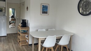 Cuina moderna amb nevera blanca, armaris de fusta i una màquina de cafè. Hi ha una zona de menjador amb taula i cadires blanques, i una catifa blava amb dibuixos.