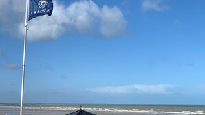 Una bandera en un pal, una platja i un edifici amb teulada.