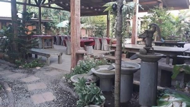 Zona patio esterna con pavimento in pietra, tavoli e sedie in legno, circondata da piante in vaso e una fontana centrale.