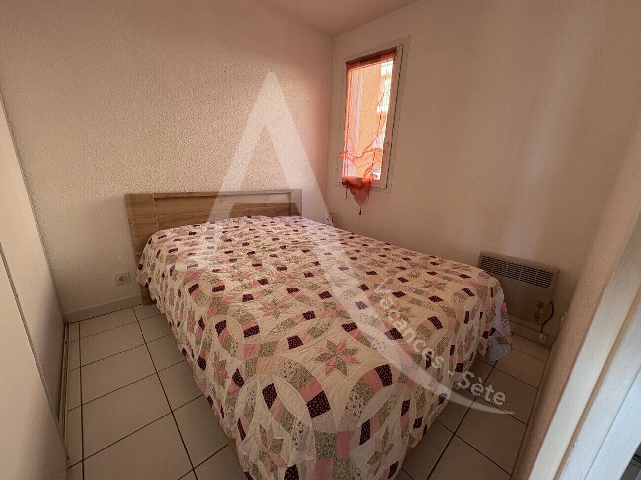 Habitació petita amb un llit individual, un panell de fusta al capçal, una finestra amb cortina vermella i un radiador a la paret.