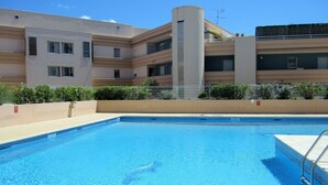 A swimming pool with a clear blue water surface, surrounded by a beige pool deck. In the background, there is a multi-story building with a flat roof and a balcony on the upper floor.