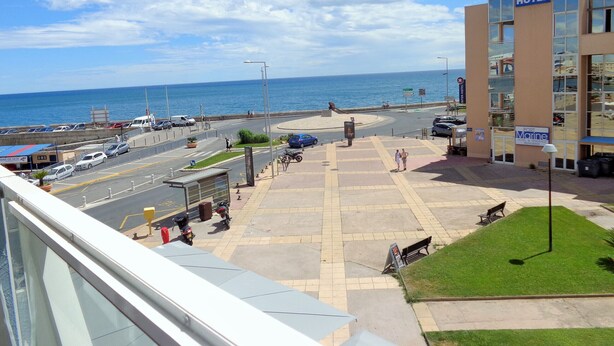 A coastal hotel with a view of the sea, a parking area, and a pedestrian walkway.