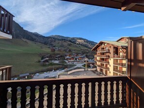 A mountain resort with wooden buildings, a balcony with a decorative railing, and a view of a valley with houses and a road.