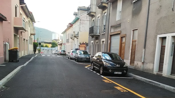 Calle bordeada por edificios de varios pisos, automóviles estacionados y un paso de peatones.