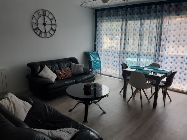 Una sala de estar con un sofá de cuero negro, una mesa redonda de centro, una mesa de comedor con sillas y un ventanal con cortinas estampadas.