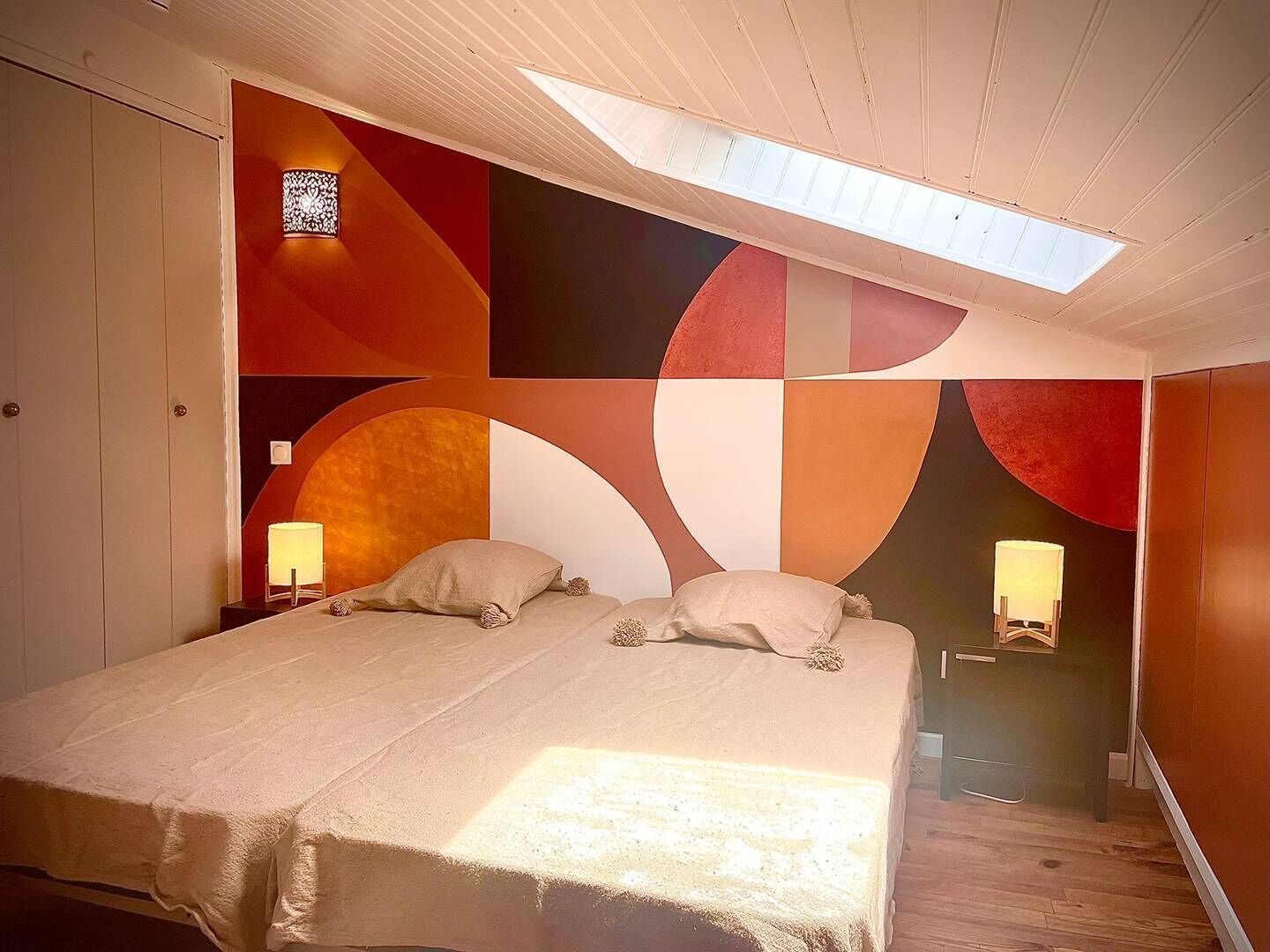 Une chambre à coucher avec un lit, deux lampes de chevet, une lucarne et un mur orné de motifs géométriques abstraits.