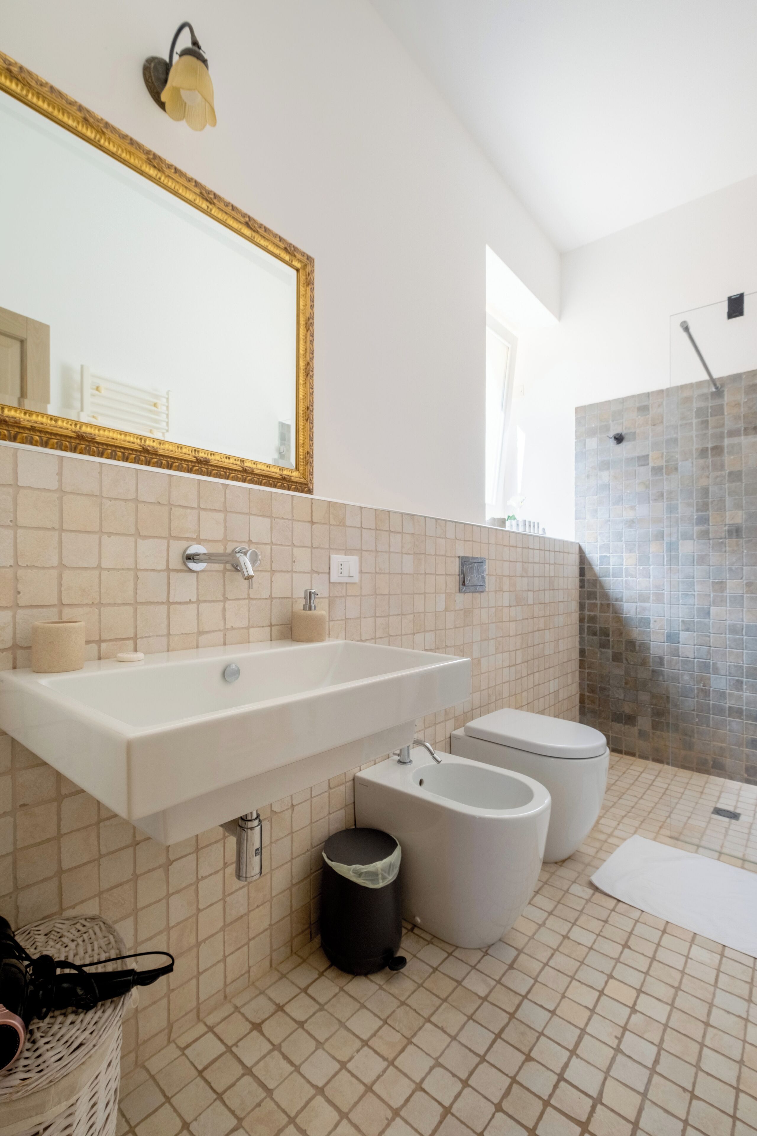 Un baño moderno con un espejo grande, un lavamanos, dos bidés, un cesto de basura y una zona de ducha.