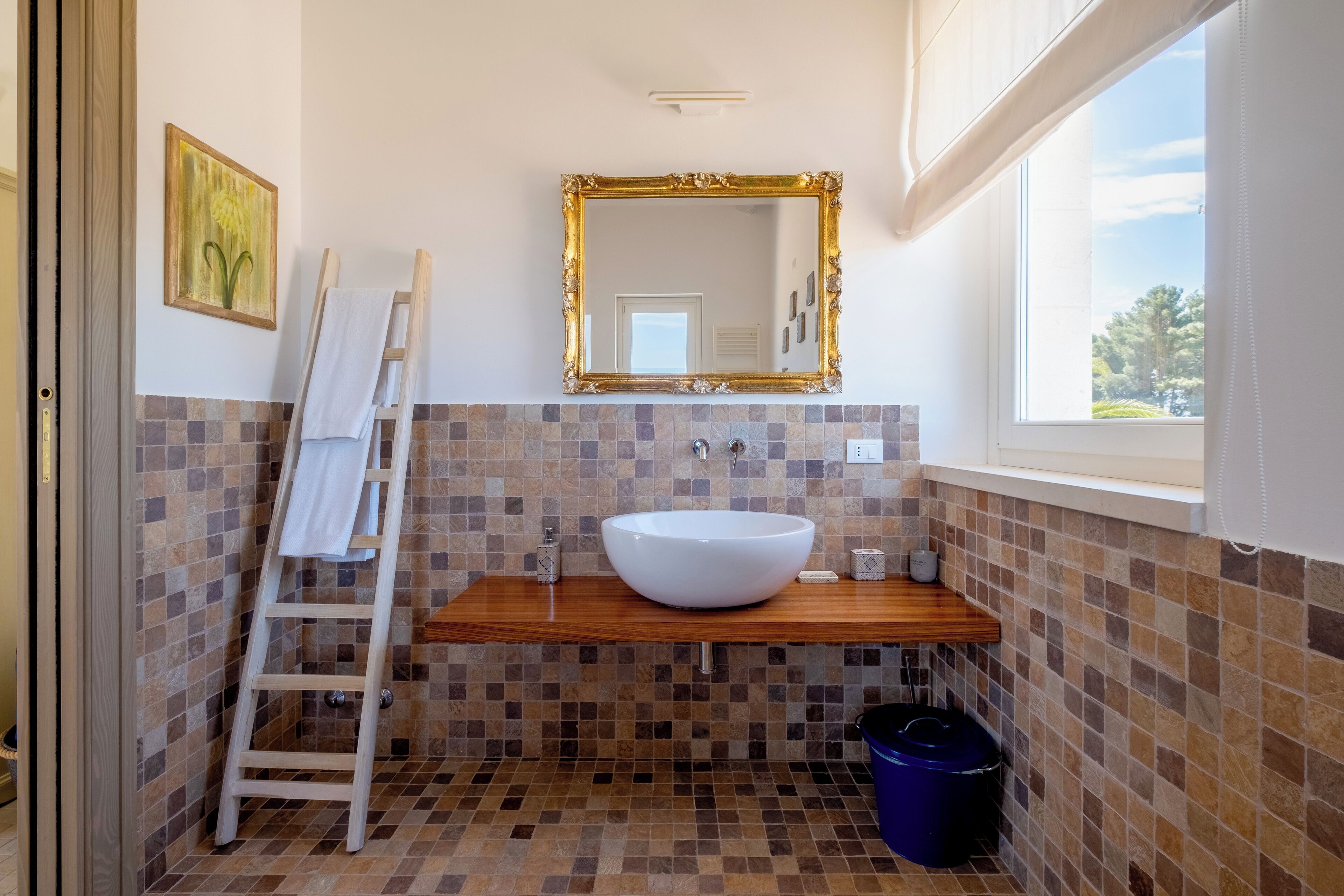 Een badkamer met een houten wastafelmeubel, een witte wastafel, een spiegel met gouden lijst, een ladder en een blauwe prullenbak.