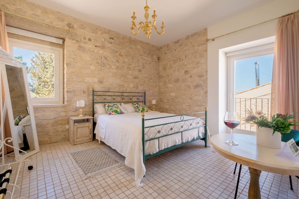 A bedroom with a stone wall, a bed with a white bedspread, a bedside table with a lamp, a round table with a glass of wine, and a window with a view of a building.