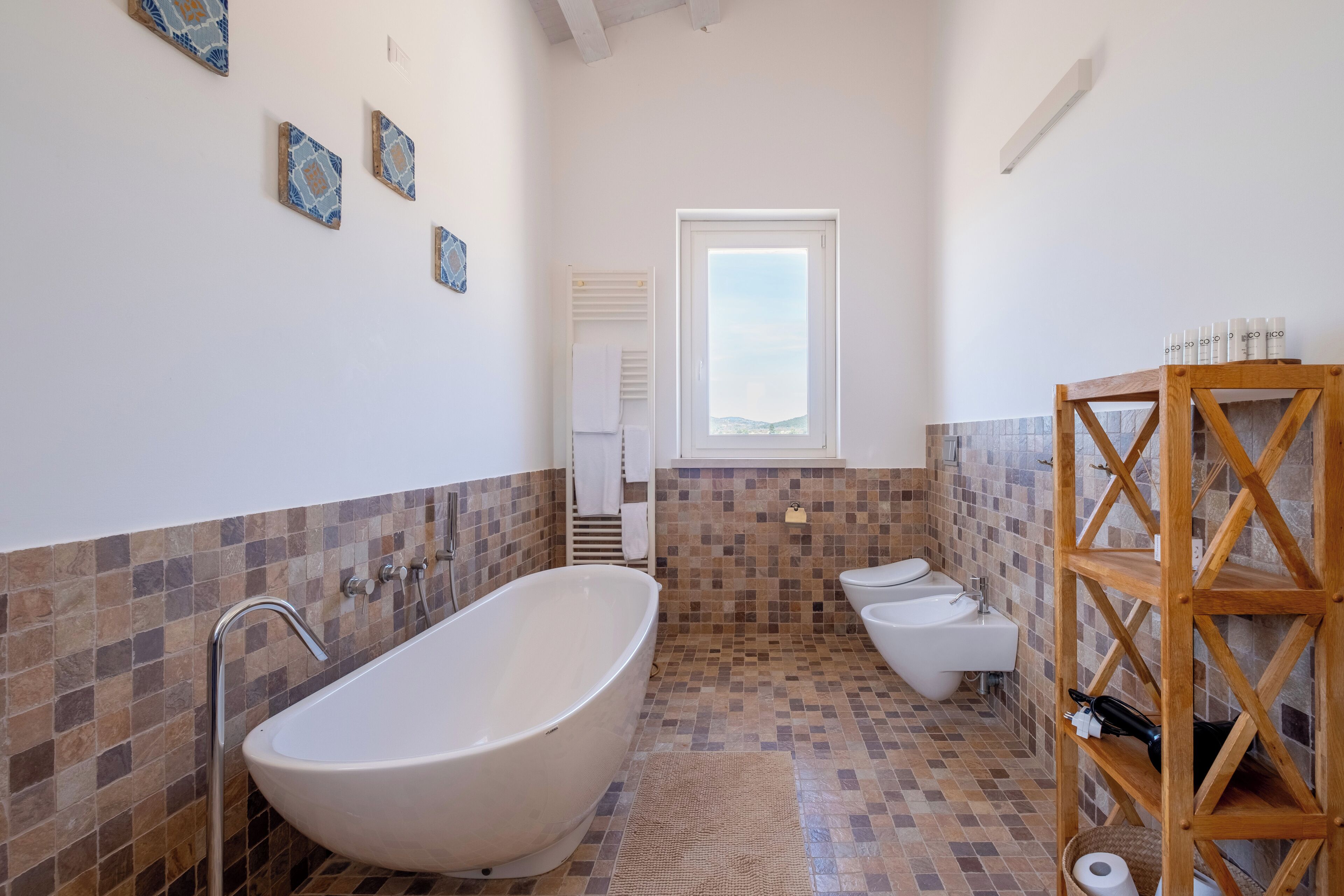 A bathroom with a freestanding bathtub, a toilet, and a bidet. There is a wooden shelf with toiletries and a window providing natural light.