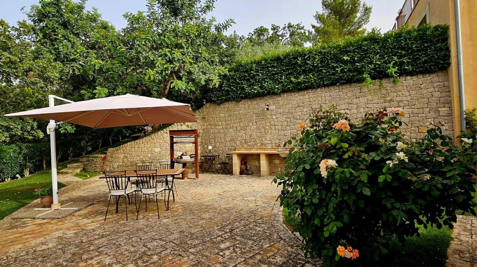 Eine Terrasse im Freien mit Steinboden, einem Tisch, Stühlen, einem großen Sonnenschirm und einer Steinwand mit Grünpflanzen.