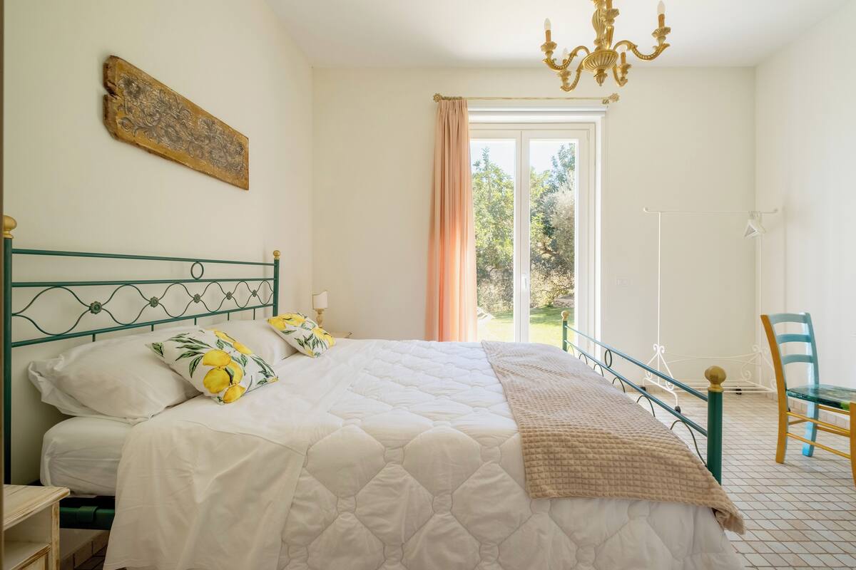 A bedroom with a bed, a chandelier, a window with curtains, and a chair.
