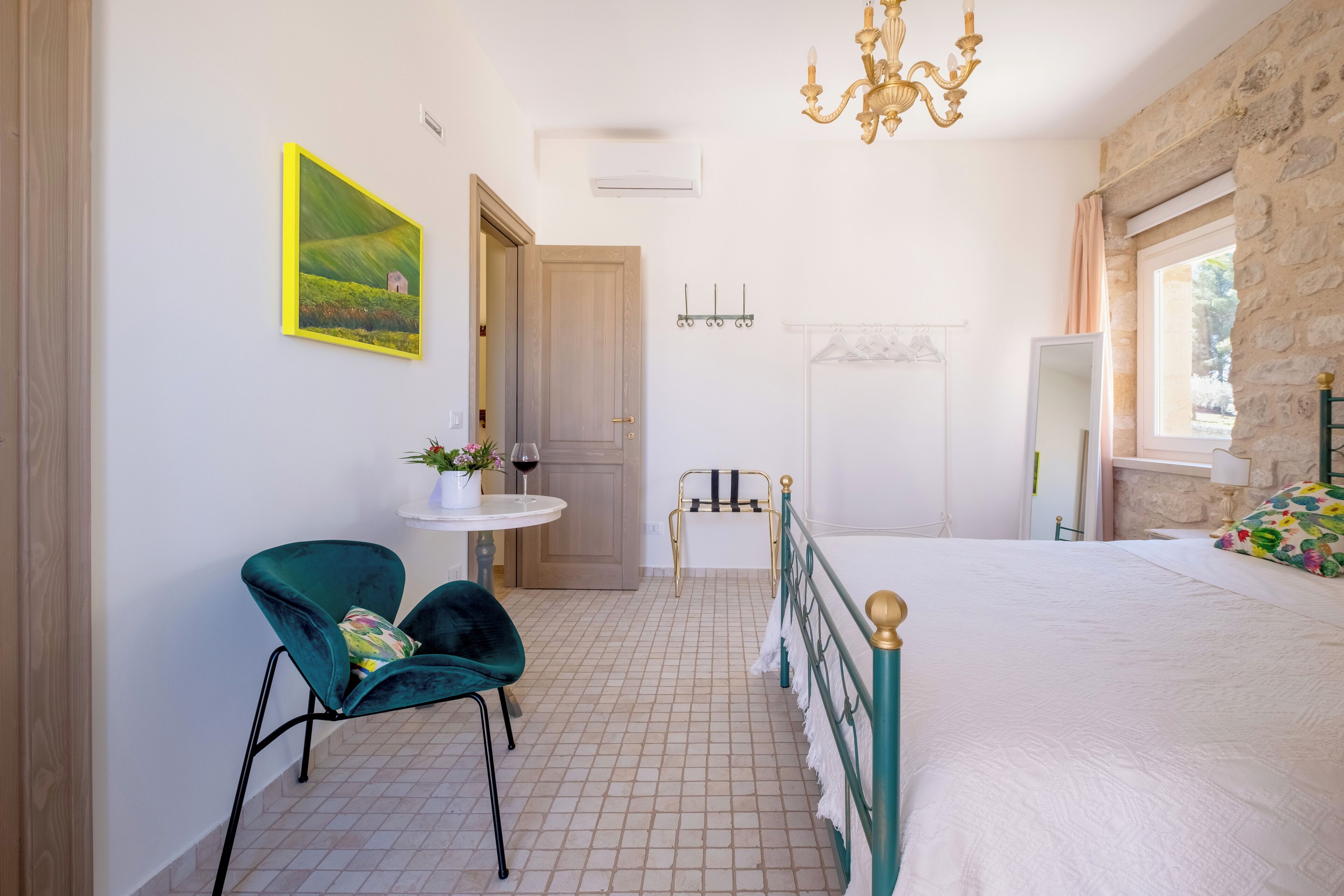 Un dormitorio con cama, una silla, una mesita con un florero y una copa de vino, un cuadro y una pared de piedra.