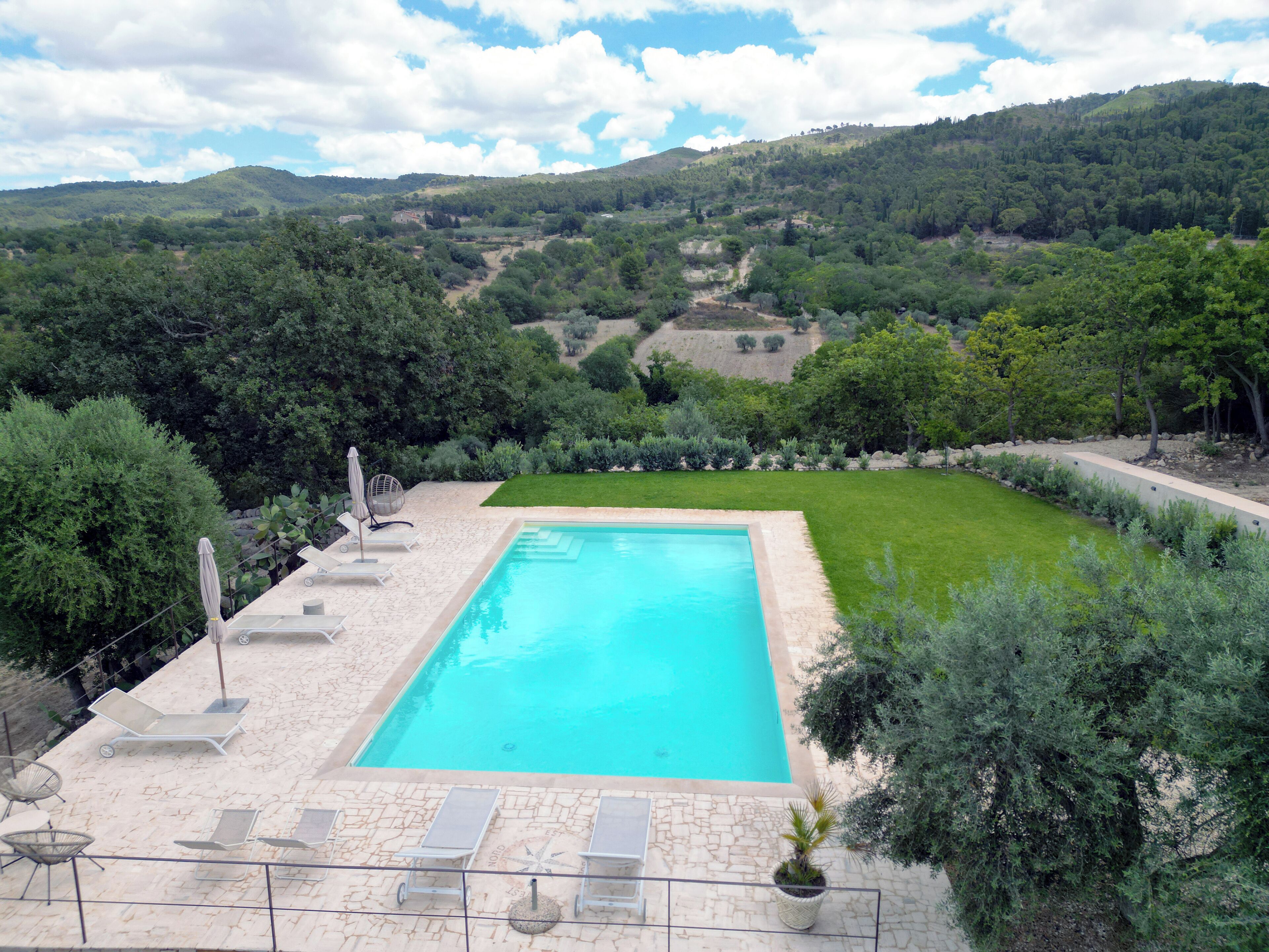 A rectangular swimming pool with a stone deck and lounge chairs, surrounded by a well-maintained lawn and trees, with hills in the background.