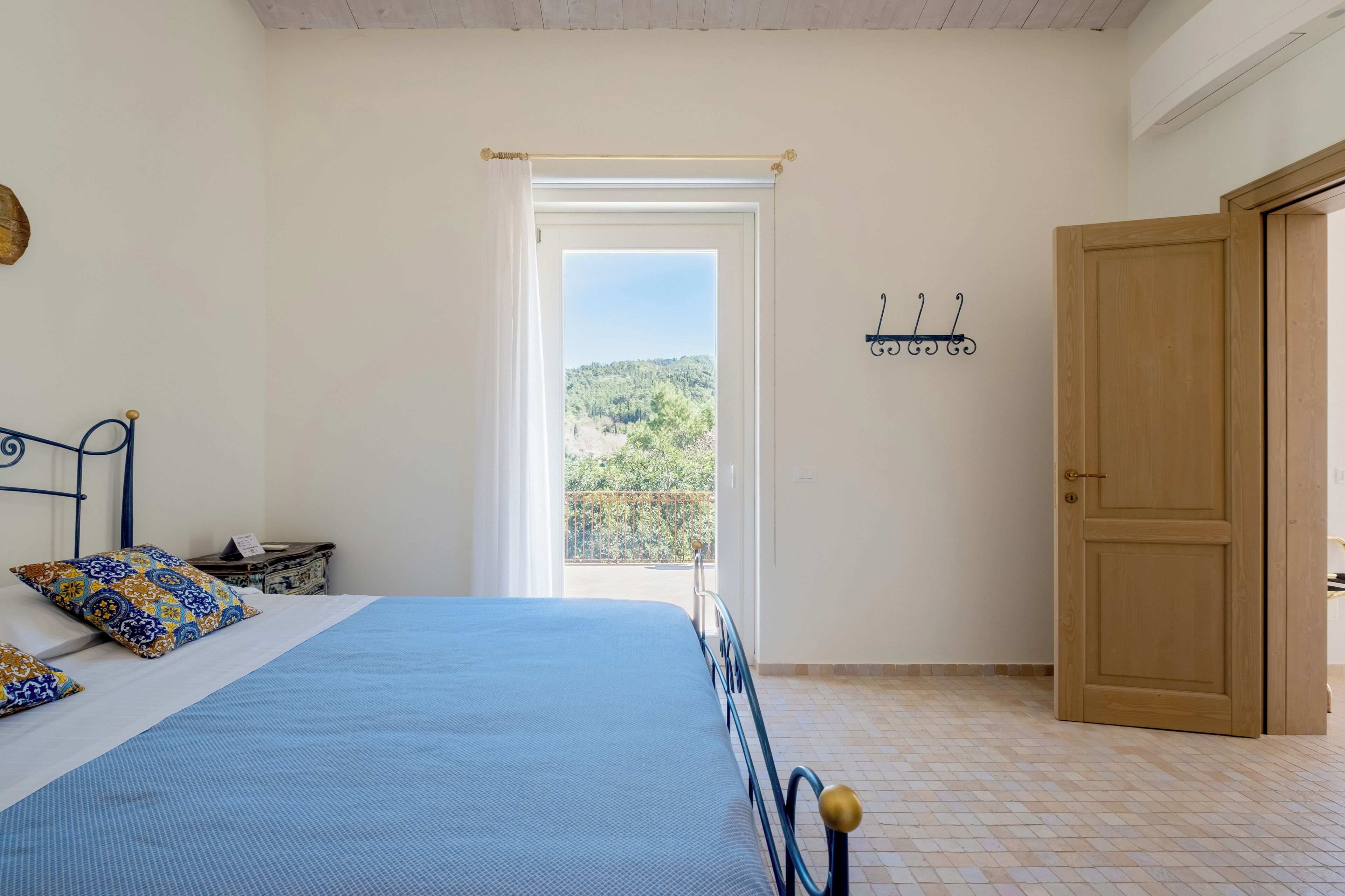 Ein Schlafzimmer mit einem Bett, einer Holztür, einem Fenster mit Blick auf Grünflächen und einem an der Wand befestigten Haken.