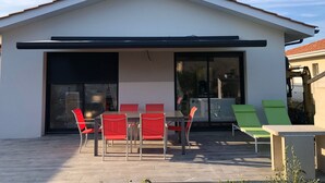 A modern house with a covered patio, a dining table with red chairs, and a green lounge chair.