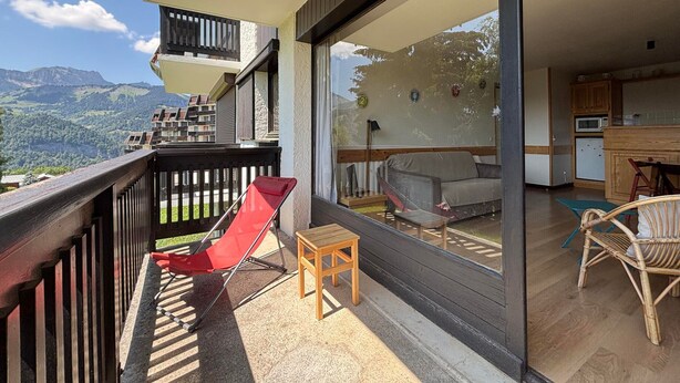 A balcony with a red chair, a small wooden table, and a view of a mountain landscape.