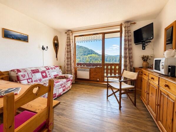 Una habitación acogedora con piso de madera, un sofá con funda estampada con motivos de corazón, una mesa de comedor con sillas, un aparador de madera, un televisor y una ventana con vista a una montaña boscosa.