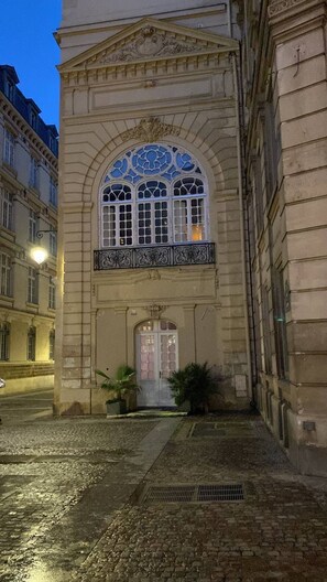Ein historisches Gebäude mit einem großen Bogenfenster, einer zentralen Tür und einem kleinen Eingangsbereich mit Topfpflanzen.