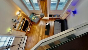 Um quarto com pé direito alto, piso de madeira, um sofá, uma mesa pequena e uma luminária.