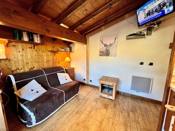 Ein gemütliches Zimmer mit einer braunen Couch, einem kleinen Tisch mit Lampe, einem Fernseher an der Wand und einem Bild von einem Reh im Rahmen.