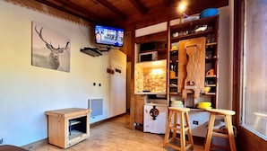 Una sala de estar acogedora con techo de madera, televisor montado, una pequeña mesa de madera y una zona de cocina con armarios y taburetes de madera.