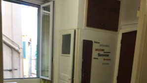 A small, compact room with a window, a door with a welcome sign, a small table with magazines, and a stool. - Studio old town 17m2, 1min from the sea (Menton)