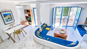 Una sala de estar moderna con un sofá curvo, una zona de comedor con una mesa y sillas blancas, y vistas al exterior a través de amplios ventanales.