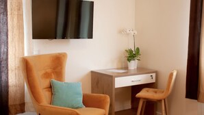 A modern living room with a large orange armchair, a smaller chair, a wooden desk with a plant, and a flat-screen TV mounted on the wall. - Hotel Isha (Frankfurt)
