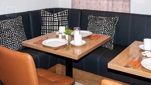 A dining area with a wooden table set for two, a black leather sofa with patterned cushions, and a brown leather chair. - Hotel Isha (Frankfurt)