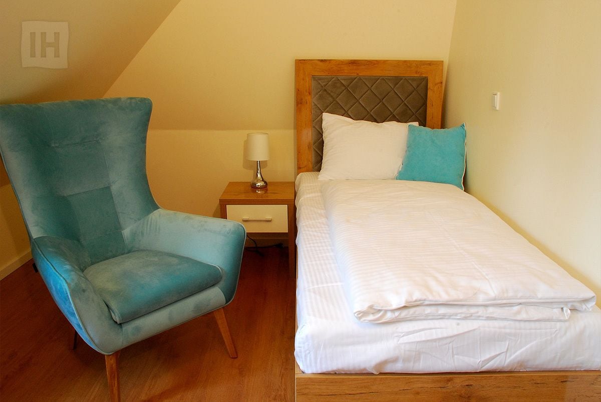 A small, neatly arranged bedroom with a single bed, a nightstand, a lamp, and a teal armchair.