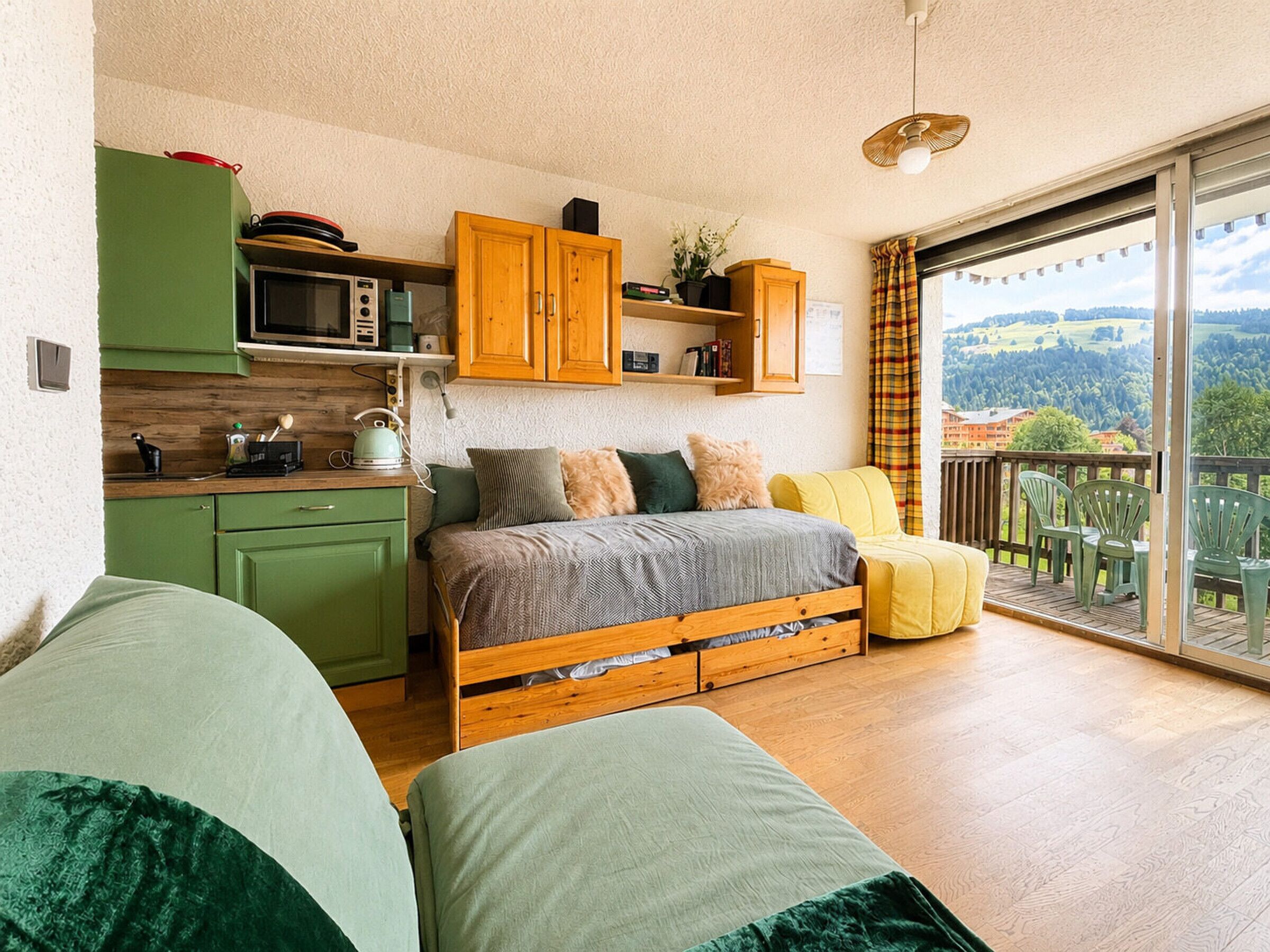 A bedroom with a bed, a green sofa, a wooden floor, a kitchen area with cabinets and appliances, and a balcony with a view.
